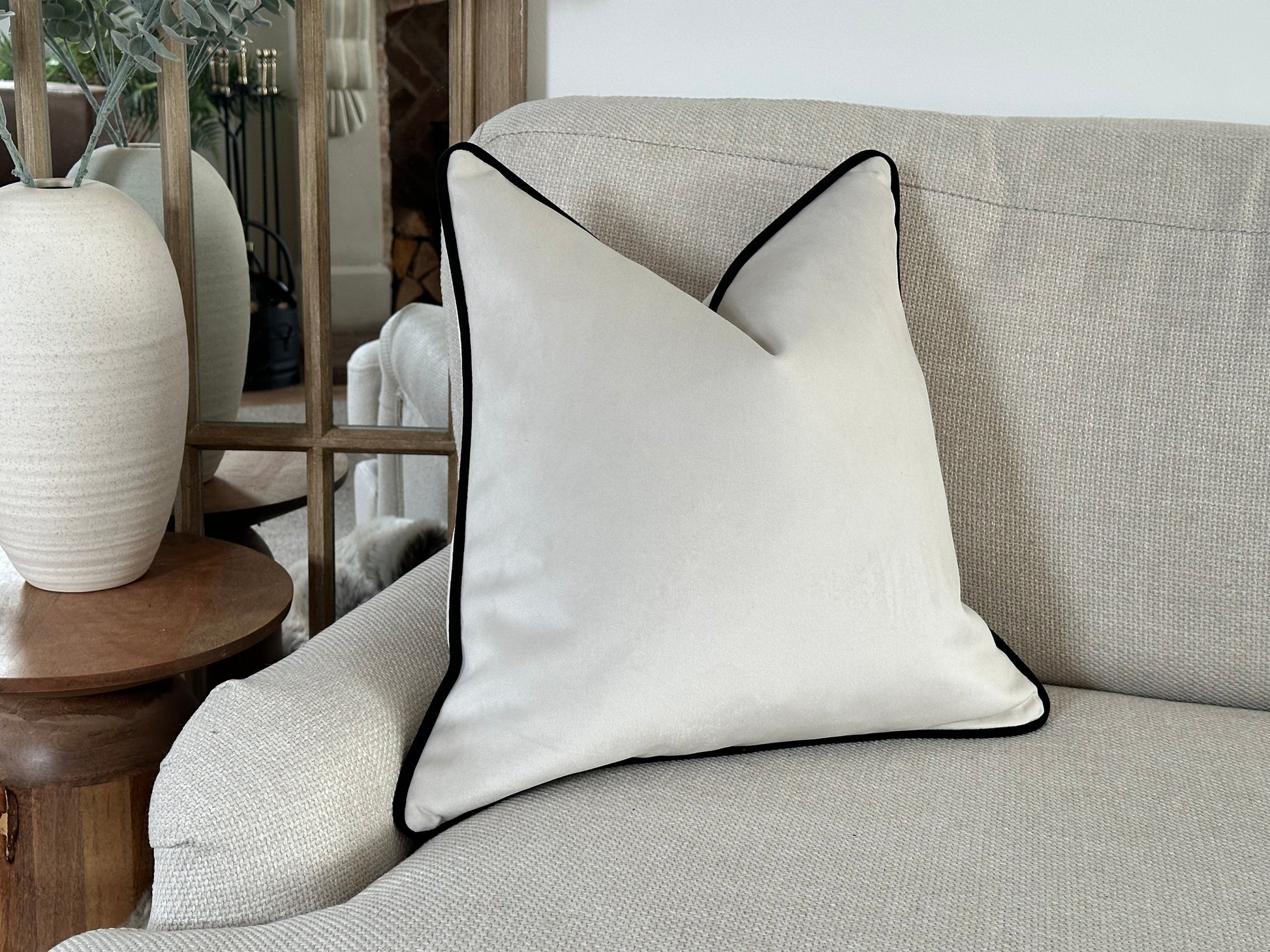 A square cushion with an off-white velvet-like fabric and contrasting black piping along the edges