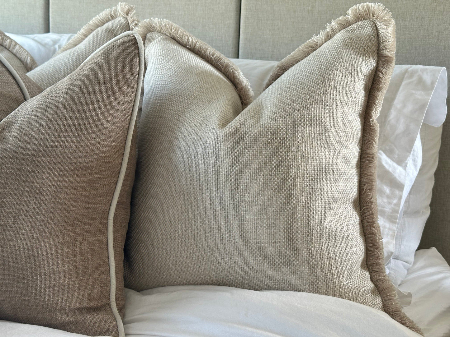 Textured beige and ivory cushion set with fringe and piping details, styled against a soft fabric headboard.