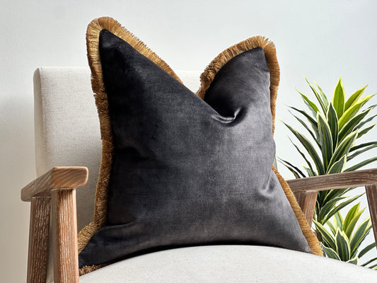 A luxury charcoal black velvet cushion with gold fringe trim placed on a neutral-toned armchair.