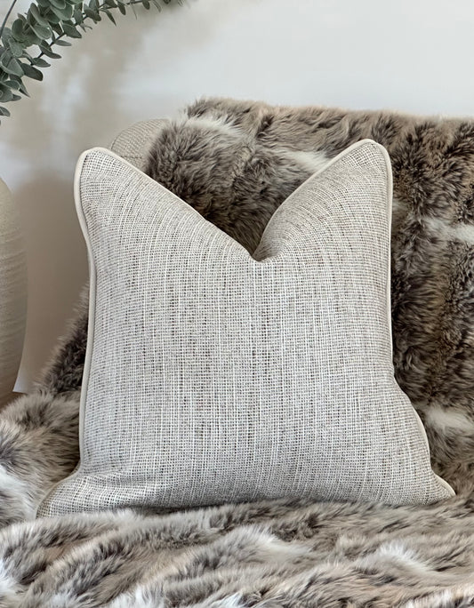 A natural textured cushion with matching piping, styled on a faux fur throw with a light armchair and ceramic vase in the background.