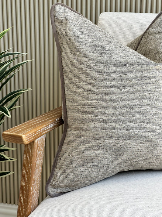 Close-up of a taupe textured cushion with deep taupe-grey piping, styled on a light neutral chair beside a plant.