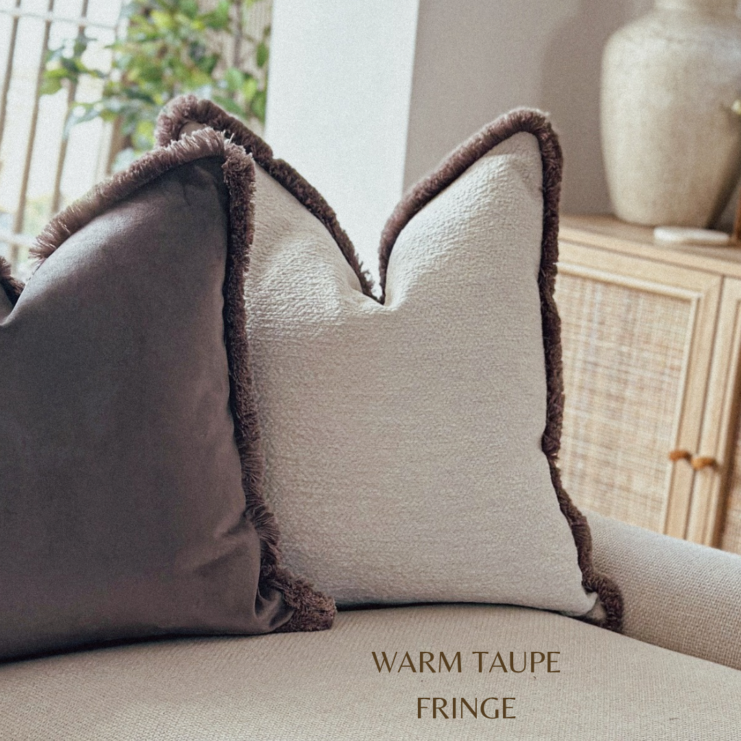 Two pillows with warm taupe fringe on a sofa, with a neutral background.