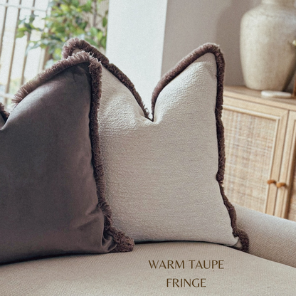 Two pillows with warm taupe fringe on a sofa, with a neutral background.