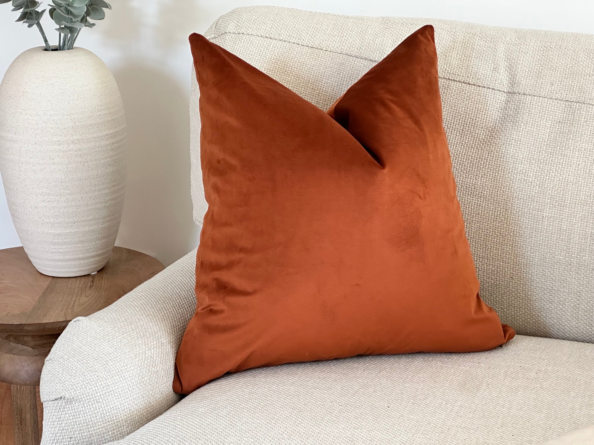 A burnt orange velvet cushion with a plain edge, styled on a light textured armchair beside a neutral ceramic vase with foliage.