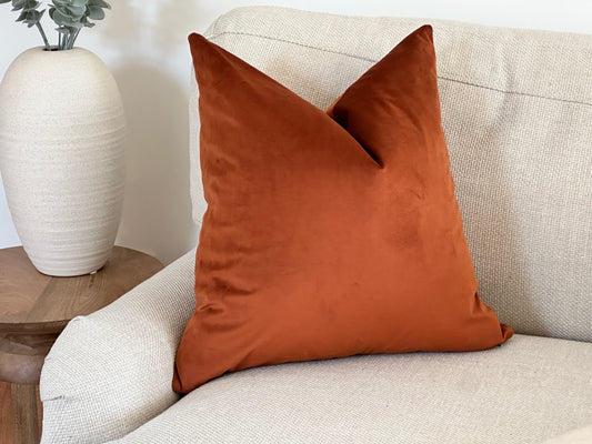 A burnt orange velvet cushion with a plain edge, styled on a light textured armchair beside a neutral ceramic vase with foliage.