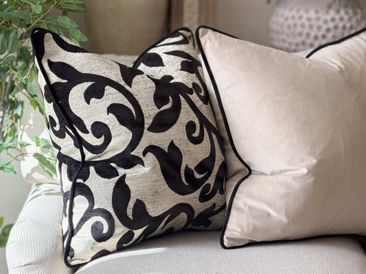 Two decorative cushions on a light-colored sofa. The front pillow has a black botanical pattern on a textured cream fabric, and the rear pillow is a solid cream velvet with black piping.