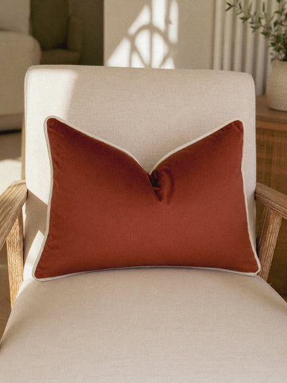 A rust-orange rectangular throw pillow with white piping, resting on a cream-colored armchair in a softly lit room.