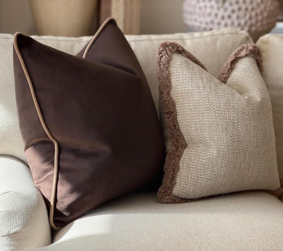 Two decorative throw pillows on a light beige sofa. One pillow is dark brown with light beige piping, and the other is beige with a brown textured trim.
