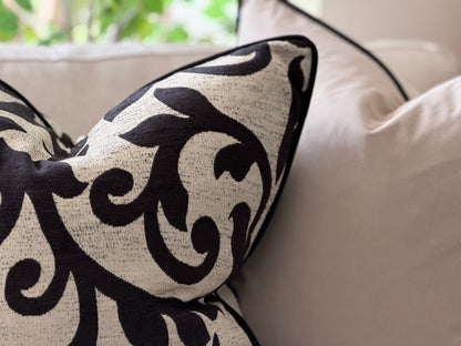A close-up of a decorative throw pillow with a black plant leafy pattern on a textured cream fabric, resting on a light beige sofa.