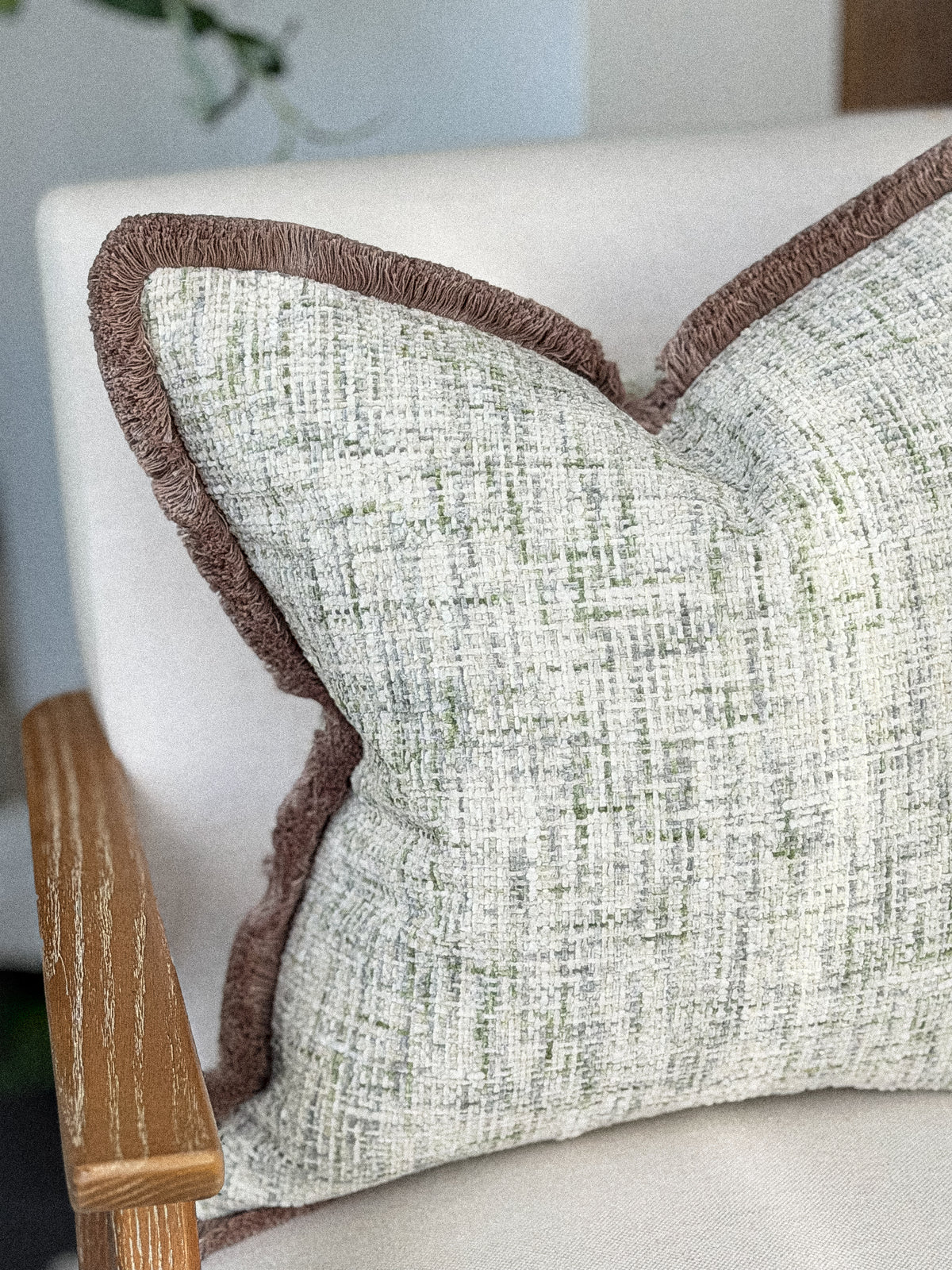A close-up of a textured throw pillow with a woven light beige and green pattern and a contrasting brown taupe fringed edge,