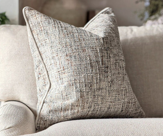 A square cushion with a textured, woven heathered pattern in beige, gray, and white and terracotta styled on a matching beige sofa.