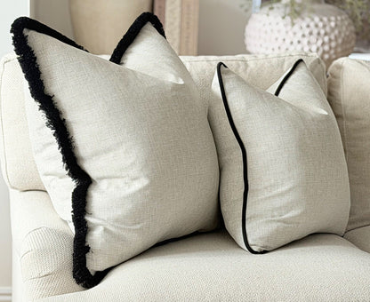 A photo of 2 cushions in a natural linen like fabric one has a black fringe trim and the other is finished with a black velvet piping.