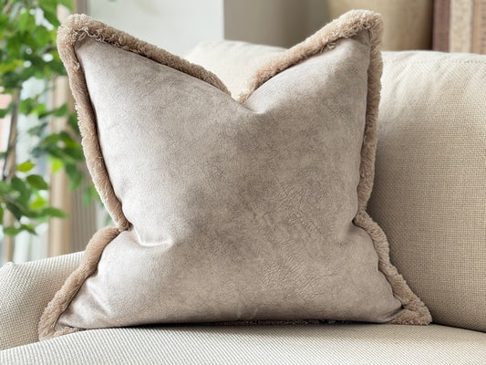 A square cushion in a soft stone ivory velvet-like fabric with a complementary trim, resting on a light beige sofa in a bright, modern living room setting.