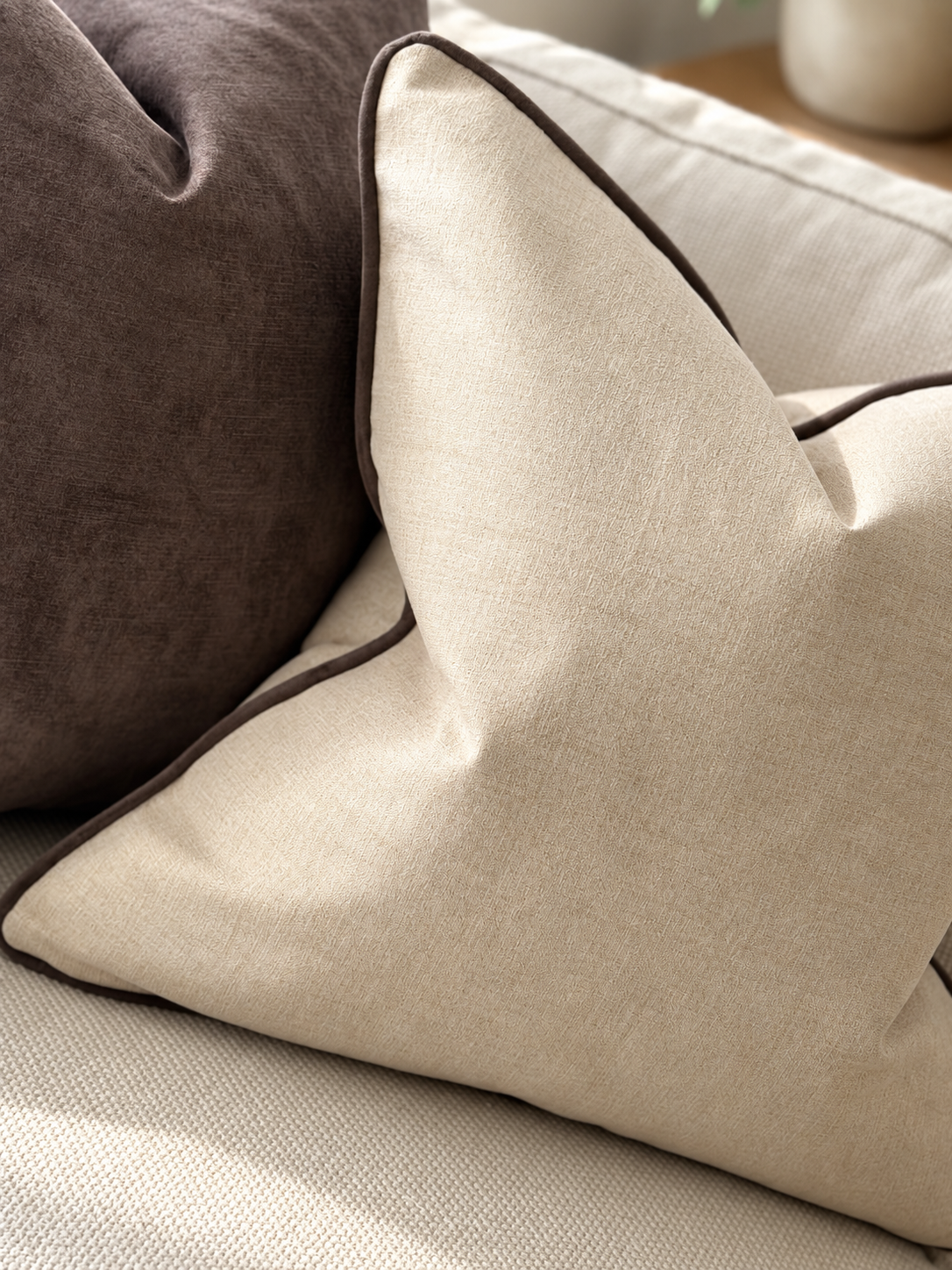 A close-up of a stone beige textured throw pillow with dark brown piping, resting on a light beige sofa next to a dark brown pillow.