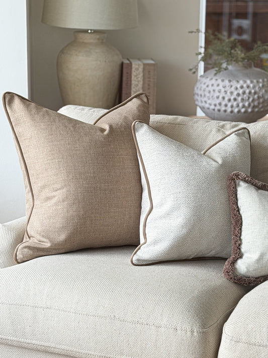 Three decorative throw pillows in neutral tones of beige, cream, and brown, styled on a light beige sofa in a cosy living room setting.
